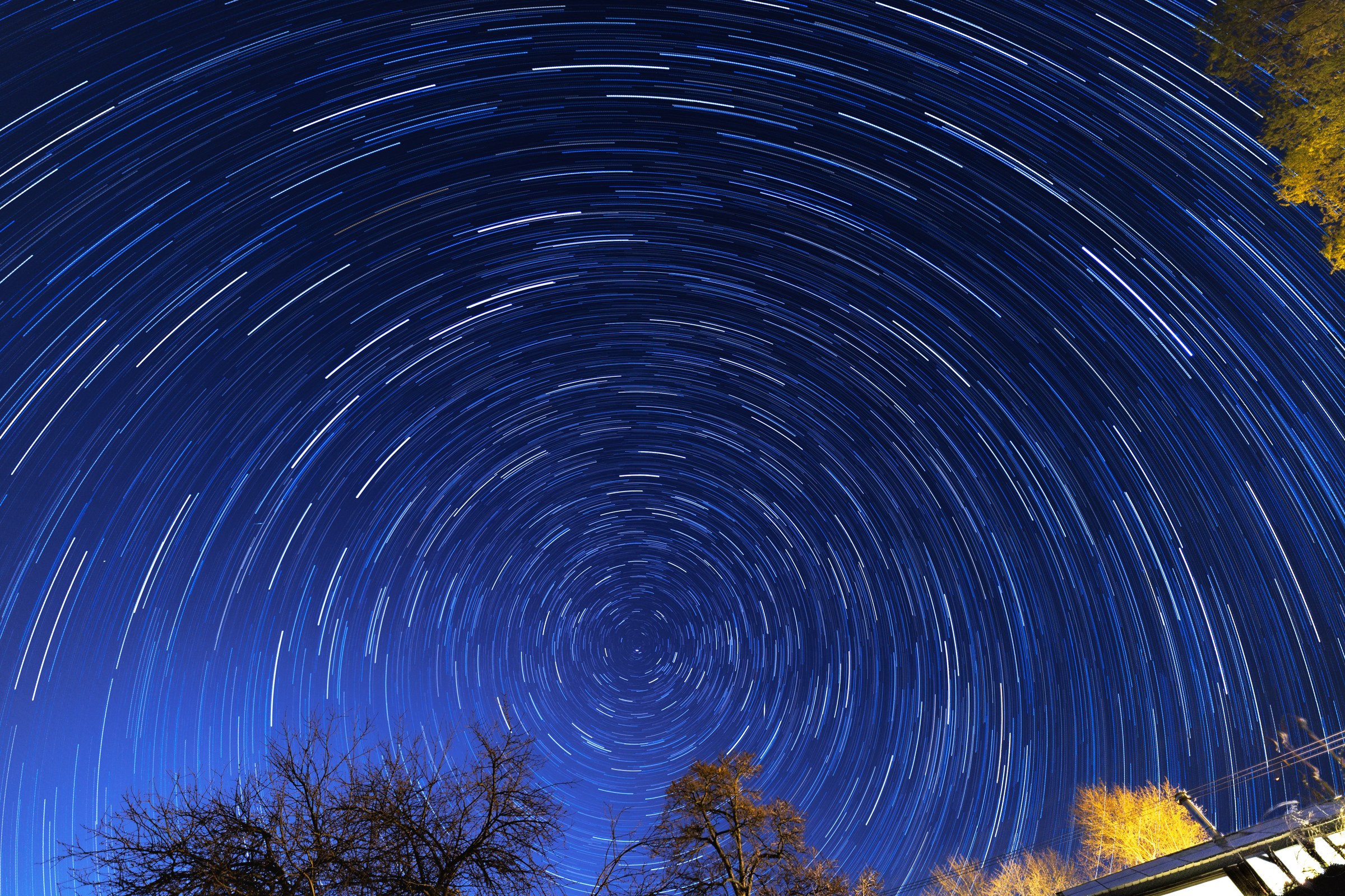 A long-exposure photograph showing circular star trails over silhouetted trees and a building.